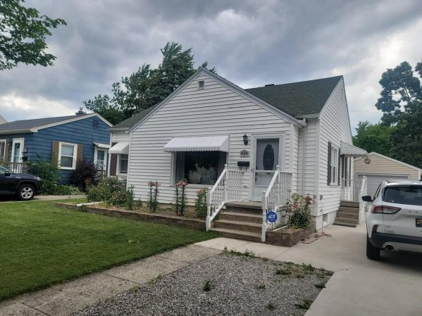 Before a custom vinyl siding remodel in Lansing, MI, showing an older white house with white vinyl siding, a small porch, and a gravel driveway. Before a custom vinyl siding remodel in Lansing, MI, showing an older white house with white vinyl siding, a small porch, and a gravel driveway.