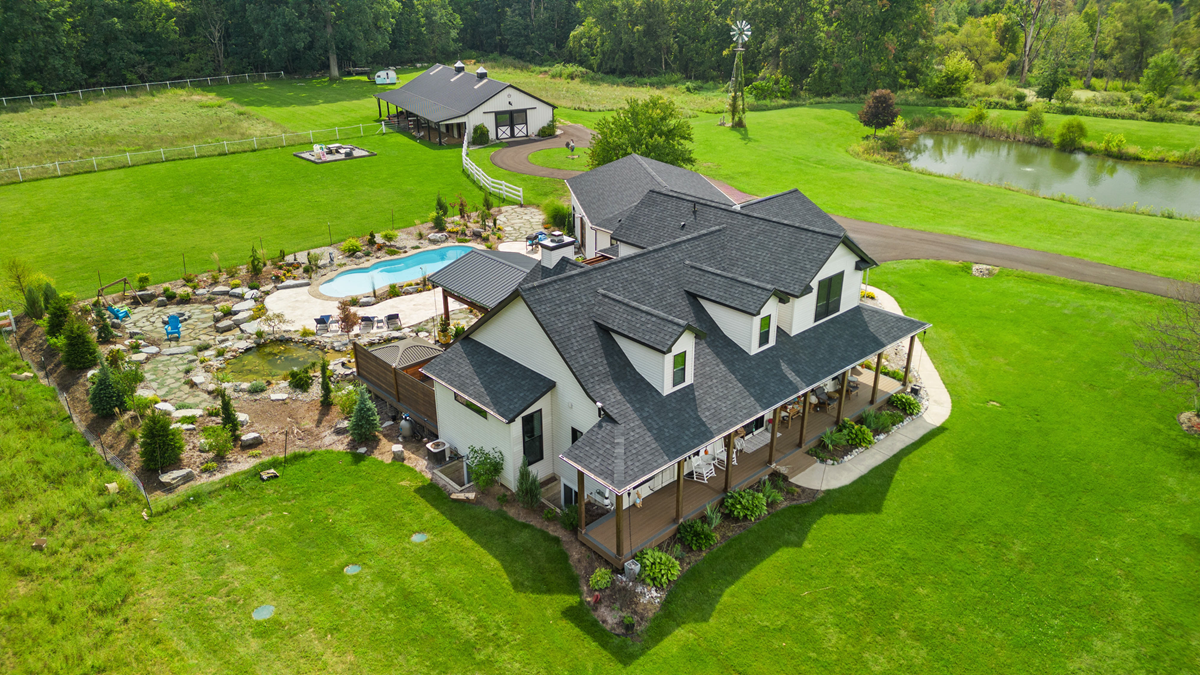 Aerial view of a white farmhouse in Holt, Michigan, with a new black roofing shingle roof, a pool, a pond, and a matching barn.