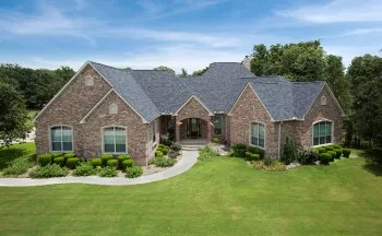 This image provides a wide, elevated view of a large, custom-built brick house in Lansing, Michigan, set against a blue sky with light clouds. The home's most prominent feature is its expansive, complex roof, which has been recently installed with new 