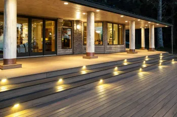This image captures a stunning TimberTech composite deck in Lansing, Michigan, showcased at dusk to highlight its sophisticated built-in lighting system. The warm, lighted step risers span the entire width of the expansive brown deck, providing both safety and a luxurious ambiance. This multi-level composite deck leads up to a covered patio with large glass doors, thick support columns, and recessed ceiling lights, all reflecting off the deck's wood-grain texture.