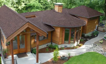 This high-angle shot showcases a beautiful, custom-designed house in Lansing, Michigan, featuring a new installation of high-quality brown roofing shingles. The dimensional asphalt shingles perfectly complement the home's warm, natural aesthetic, which includes natural wood siding and stone veneer accents. The complex roofline, with its various gables and a unique curved section, is expertly covered by the brown roofing shingles. The home is surrounded by lush trees and new landscaping, with a concrete walkway winding through the yard.